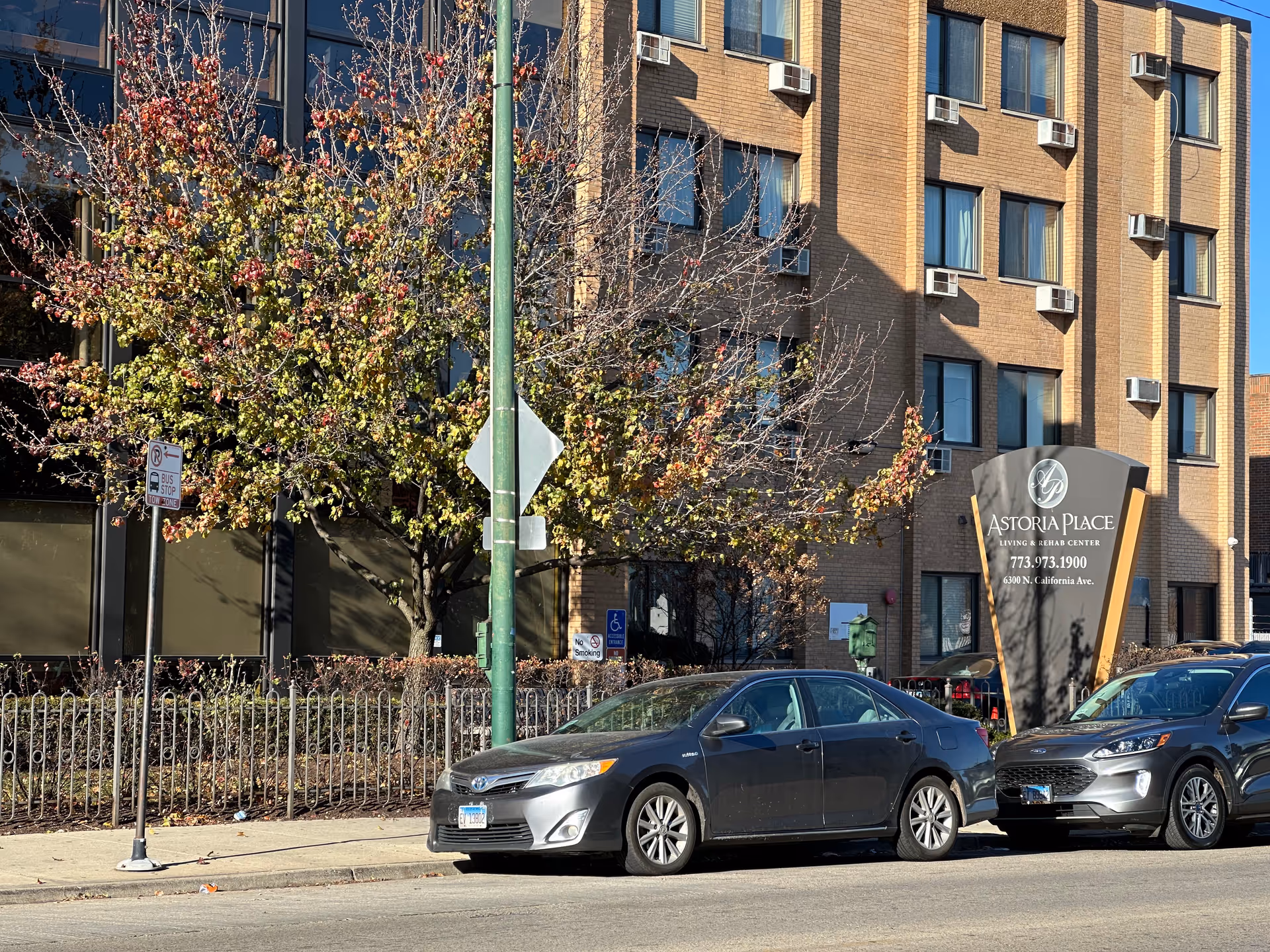 Brick facade of Astoria Place living and rehab center with its entrance sign, parked cars, and a tree along the sidewalk.