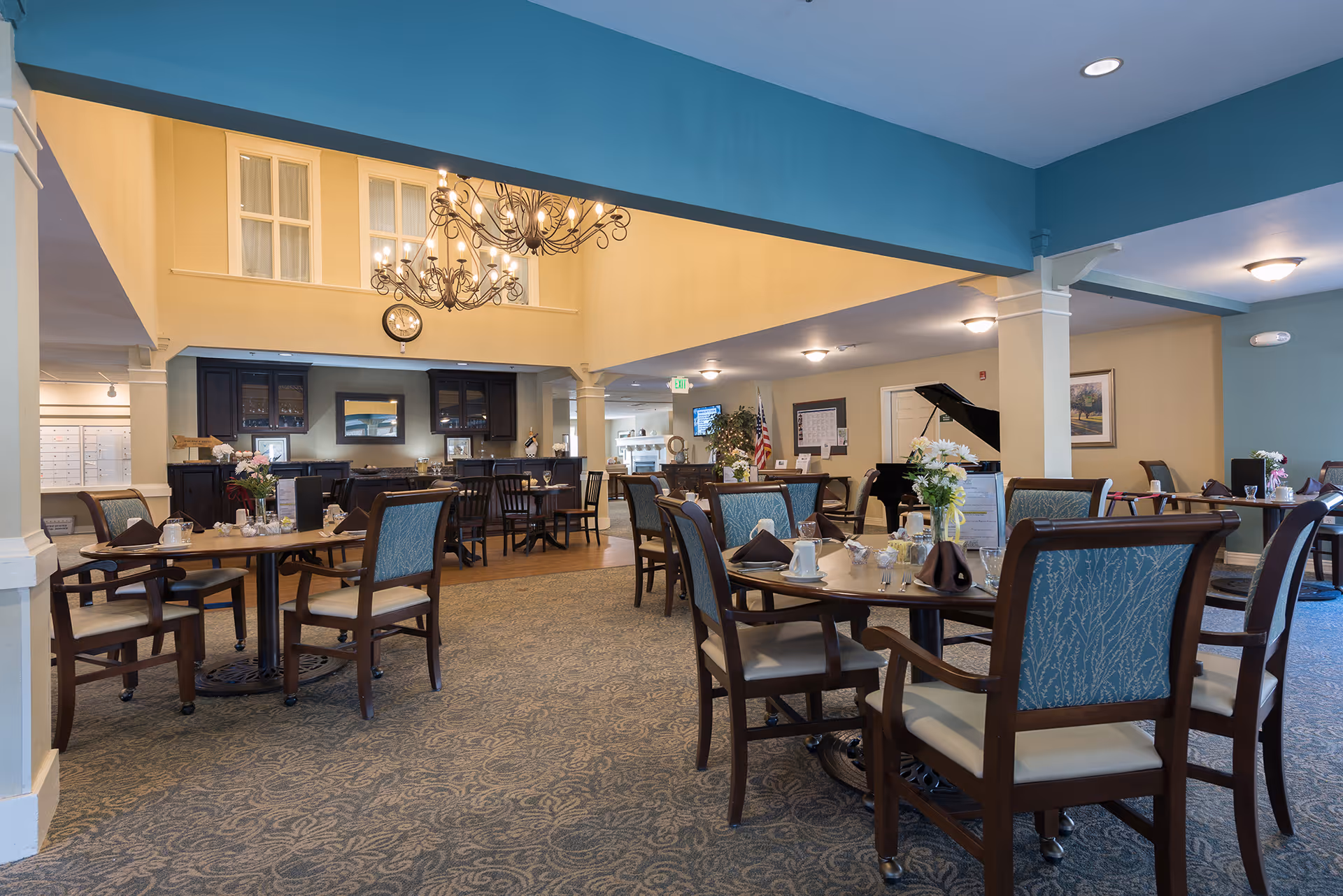 A spacious dining room in a senior living facility with multiple wooden tables and chairs arranged neatly. Each table is set with napkins, cups, and small flower arrangements. The room features a high ceiling with a large chandelier, a carpeted floor, and a piano in the background. The walls are painted in soft yellow and blue tones, creating a warm and inviting atmosphere.