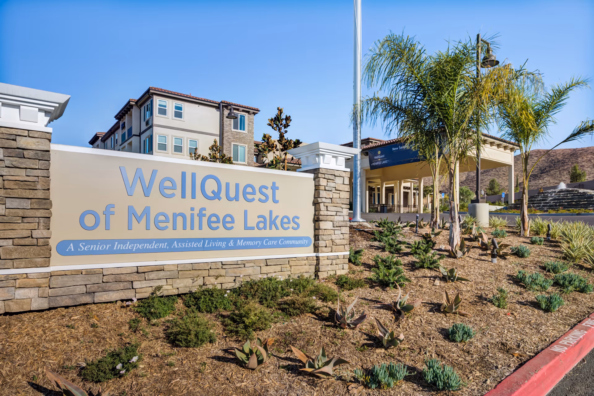 Entrance sign for WellQuest of Menifee Lakes, a senior independent, assisted living, and memory care community, with landscaped plants and a multi-story building in the background under a clear blue sky.