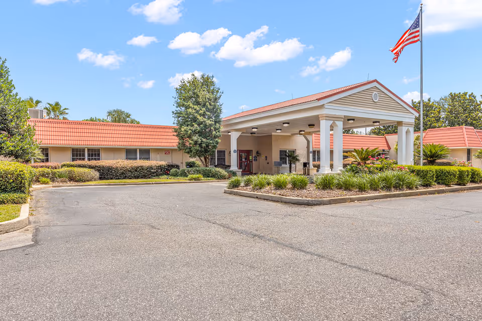 Front entrance and porte-cochère of a one-story senior living building with landscaped beds and an American flag.