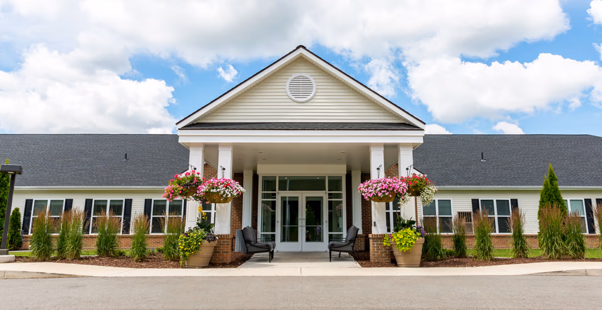 Front exterior view of a single-story building with a peaked roof, white siding, and brick accents. The entrance features a covered porch with white columns, hanging flower baskets, potted plants, and two black chairs. The sky is partly cloudy with blue patches.