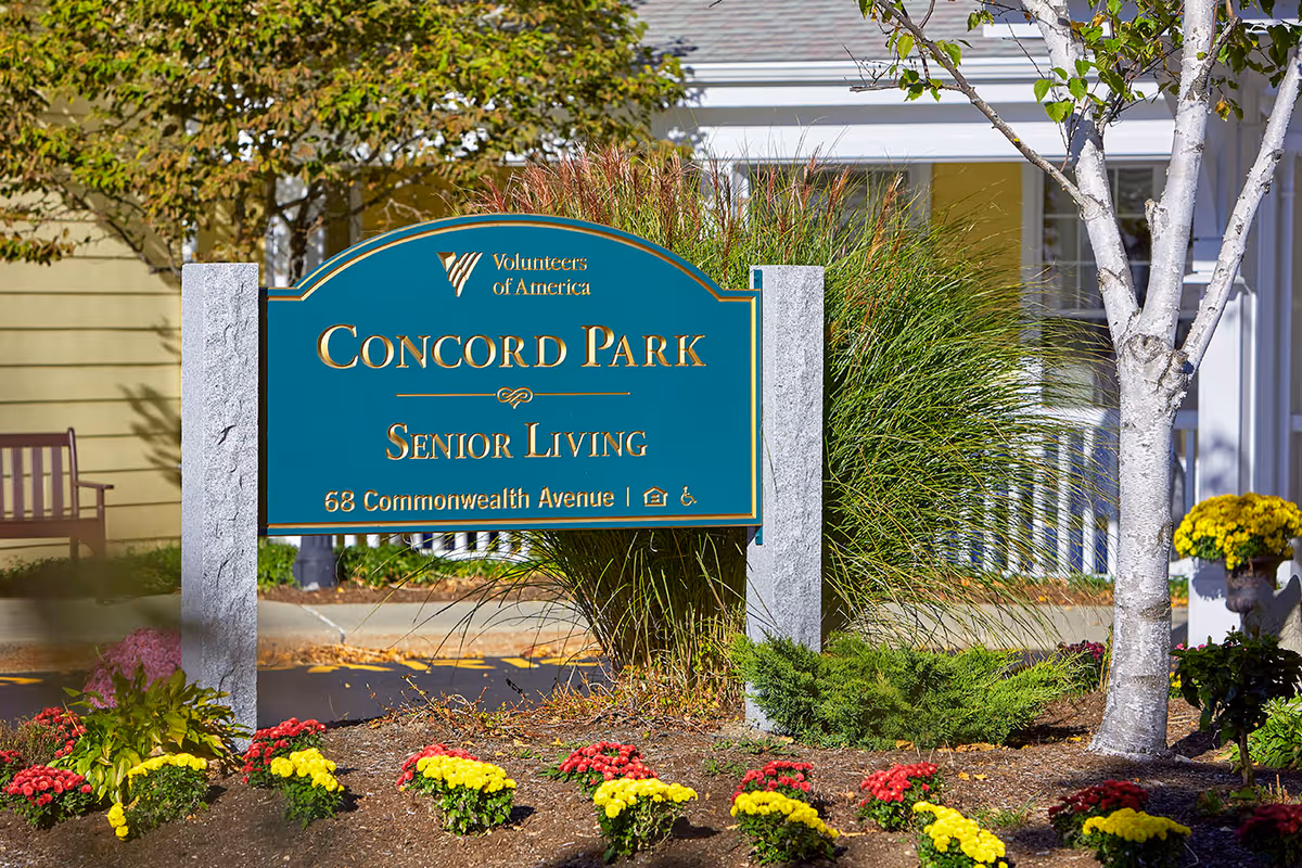 A teal entrance sign reading "Concord Park Senior Living" set in a landscaped area with flowers, trees, and a building facade behind it.