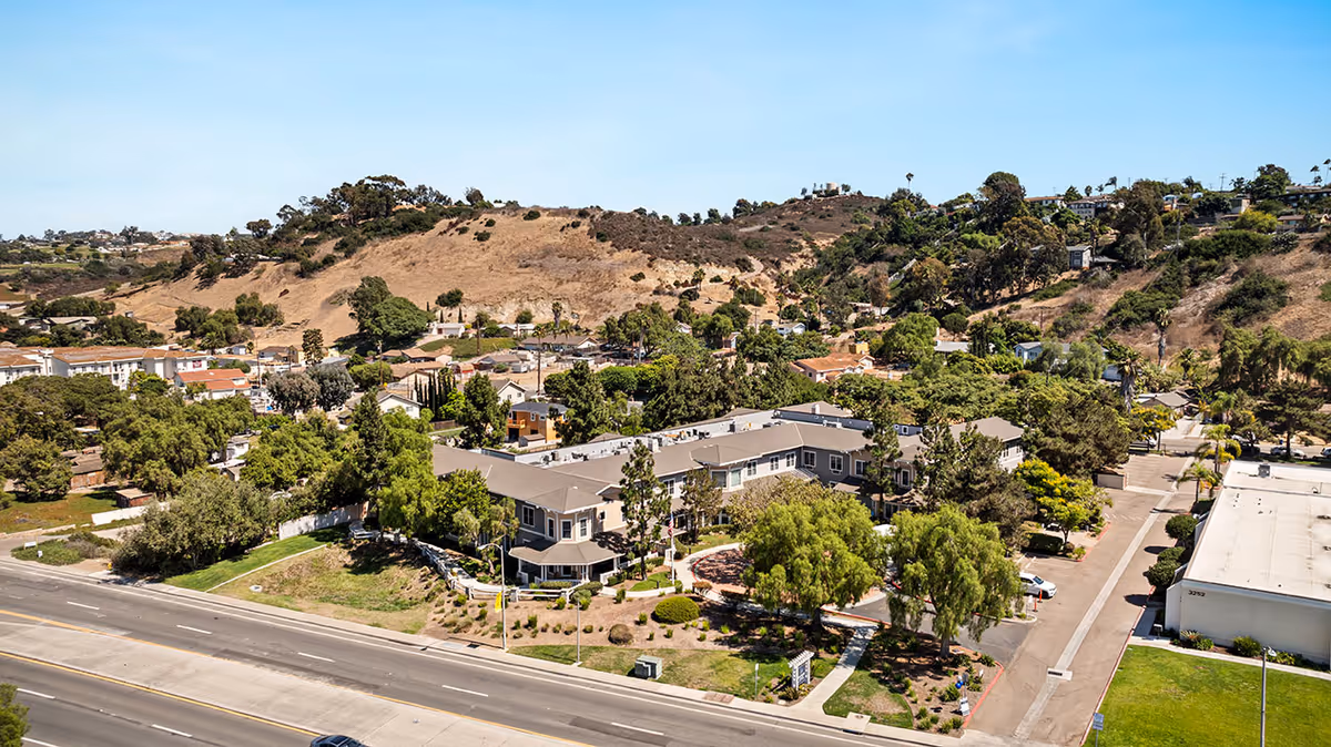 Aerial view of a senior living facility named Ivy Park at Bonita surrounded by trees and landscaping, with a large hill and residential neighborhood in the background under a clear blue sky.