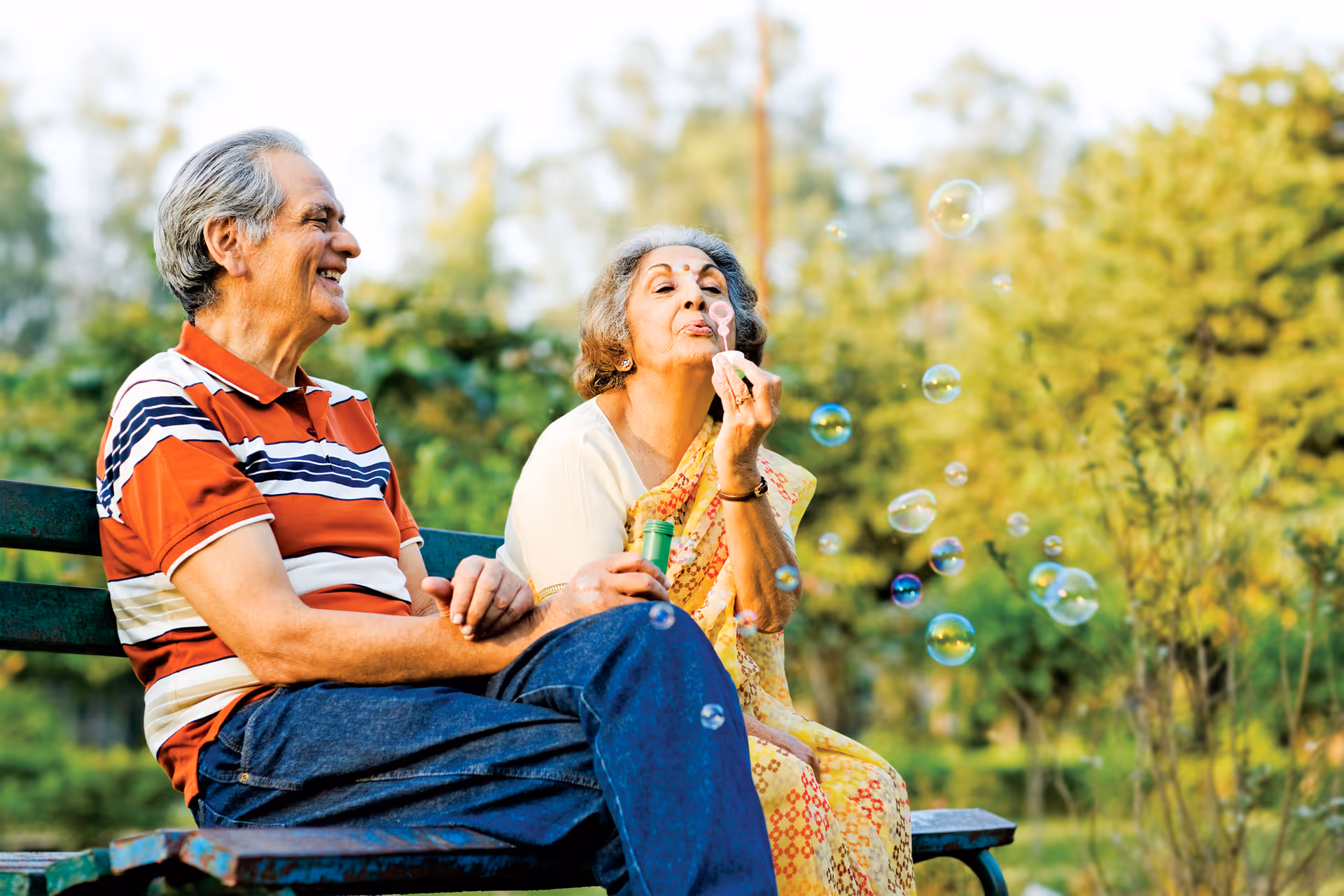 Two elderly people sit on a bench outdoors blowing bubbles.