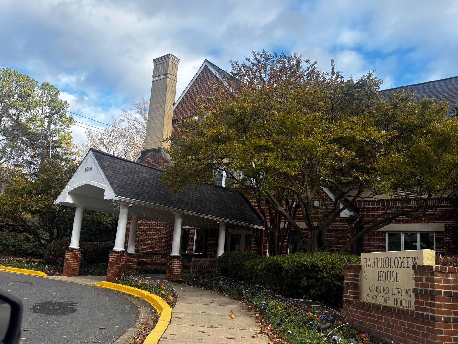 Front entrance of a brick assisted living building with a covered portico, trees, and a sign reading 'Bartholomew House'.