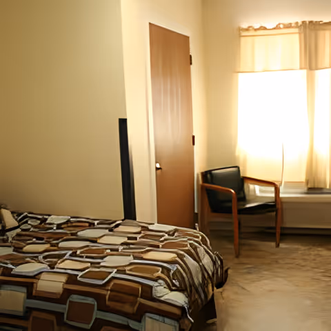 Bedroom with a patterned bedspread, a closed wooden door, and a chair by a sunlit curtained window.