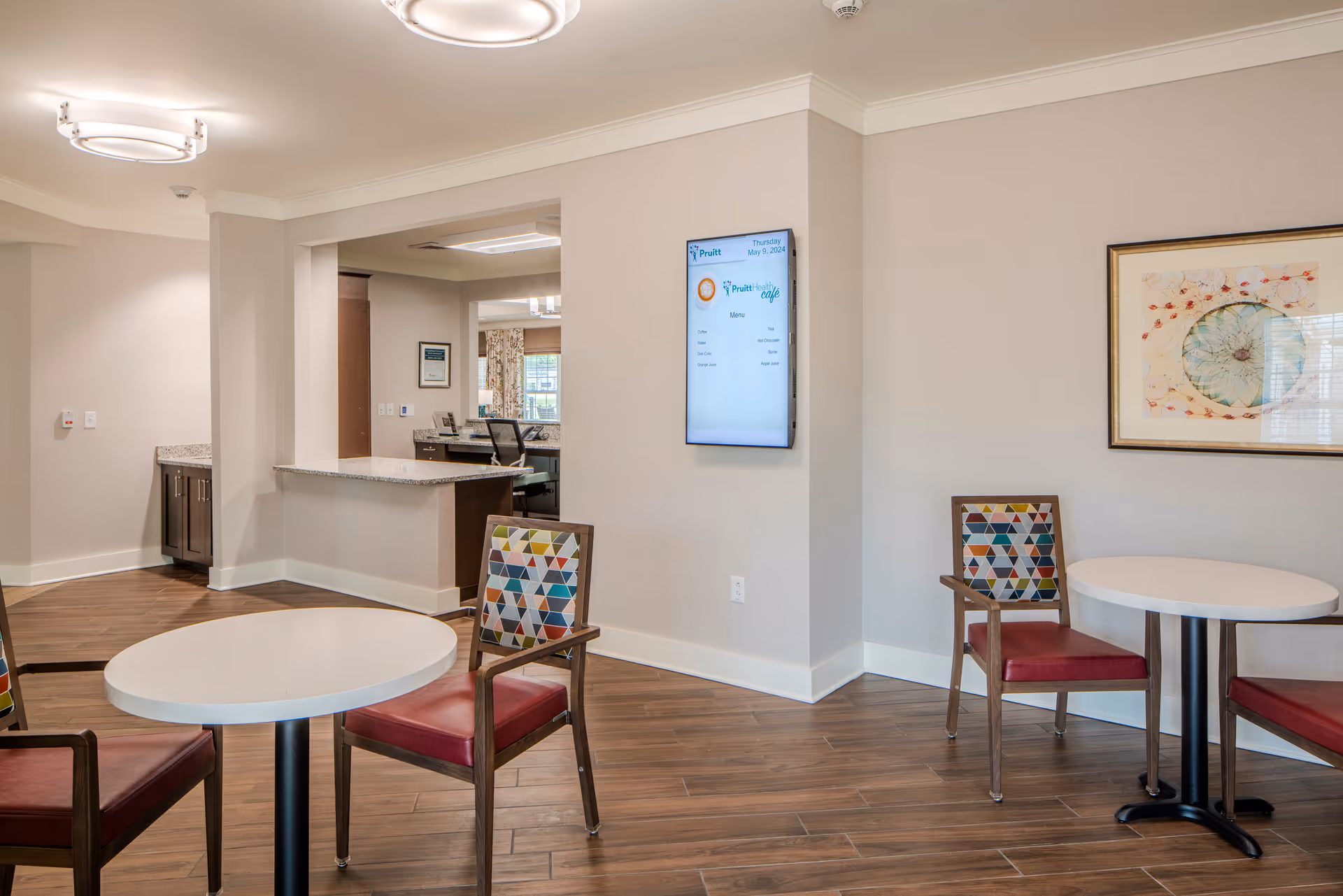 Bright lounge-dining area with round tables, colorful chairs, and a wall-mounted screen.