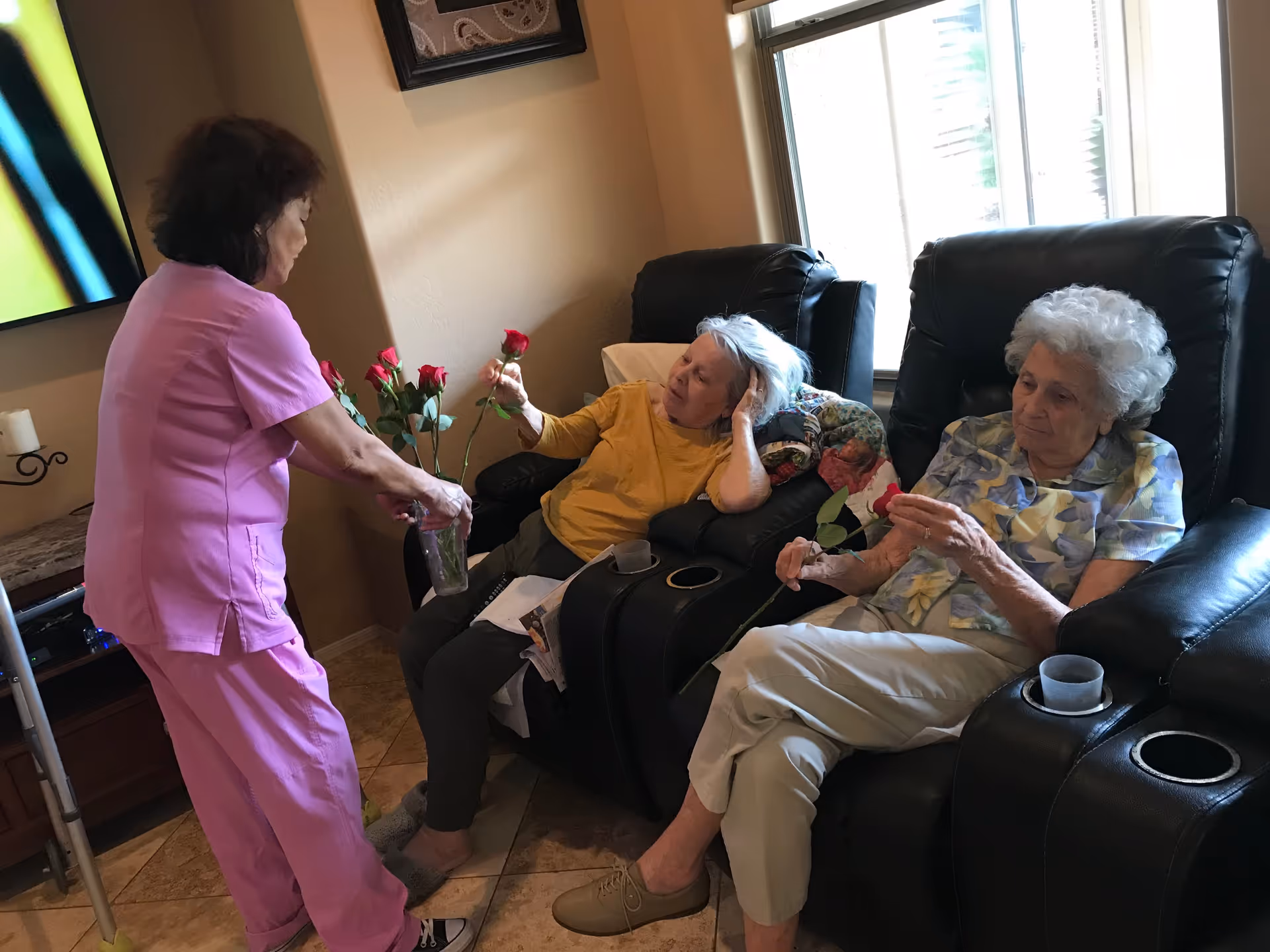 A caregiver in pink scrubs is handing out red roses to two elderly women seated in black recliner chairs in a cozy living room with a window in the background.