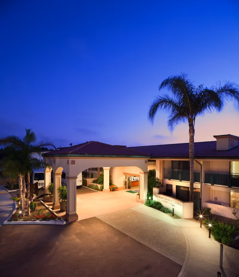 Exterior view of White Sands La Jolla facility at dusk, showing the entrance with a covered driveway, palm trees, and well-lit pathways.