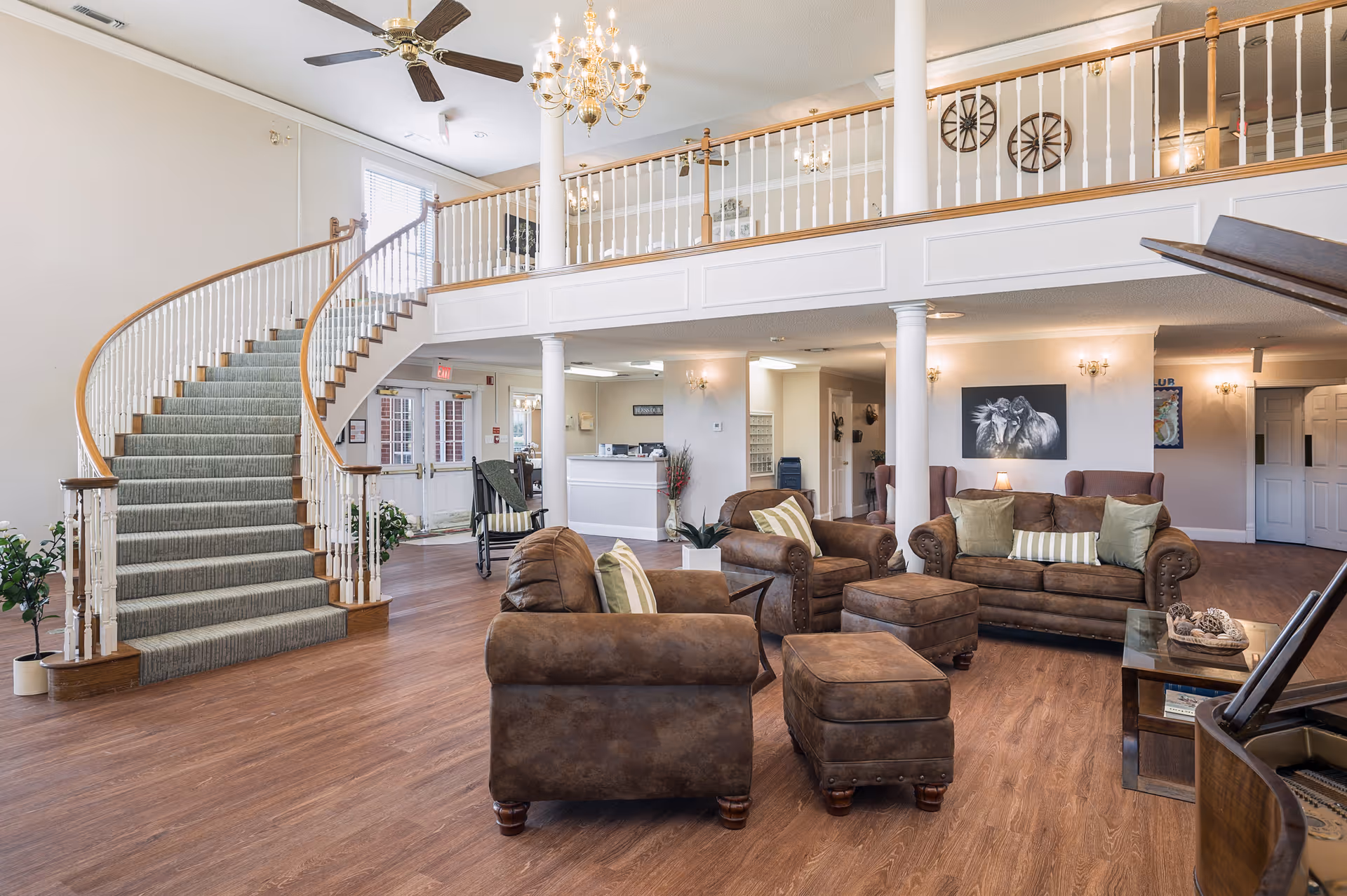 Spacious senior living facility common area with a curved staircase on the left, brown leather sofas and armchairs with green and striped pillows, a wooden floor, white pillars, a chandelier, ceiling fans, and a piano on the right side. The upper level has a white railing and decorative wagon wheels on the wall.