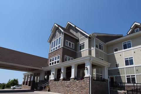 Exterior view of a multi-story senior living facility building with a covered entrance supported by white columns and stone facade under a clear blue sky.
