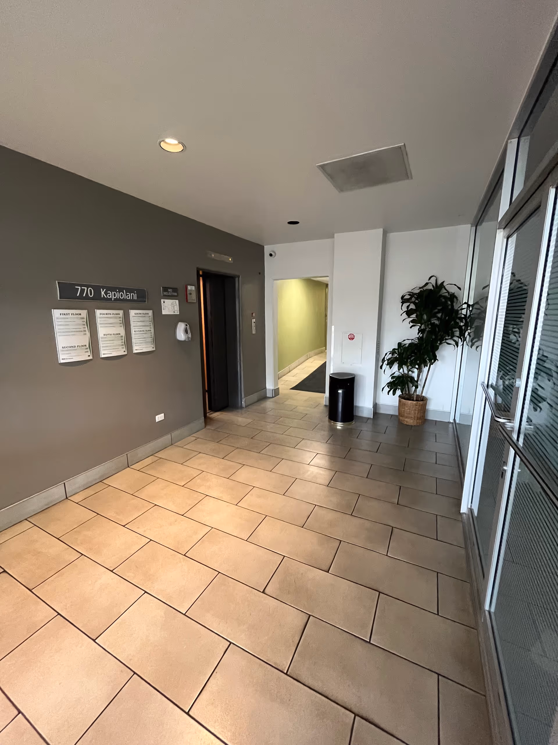 Interior corridor of a senior living facility with elevator, plants, and glass doors.