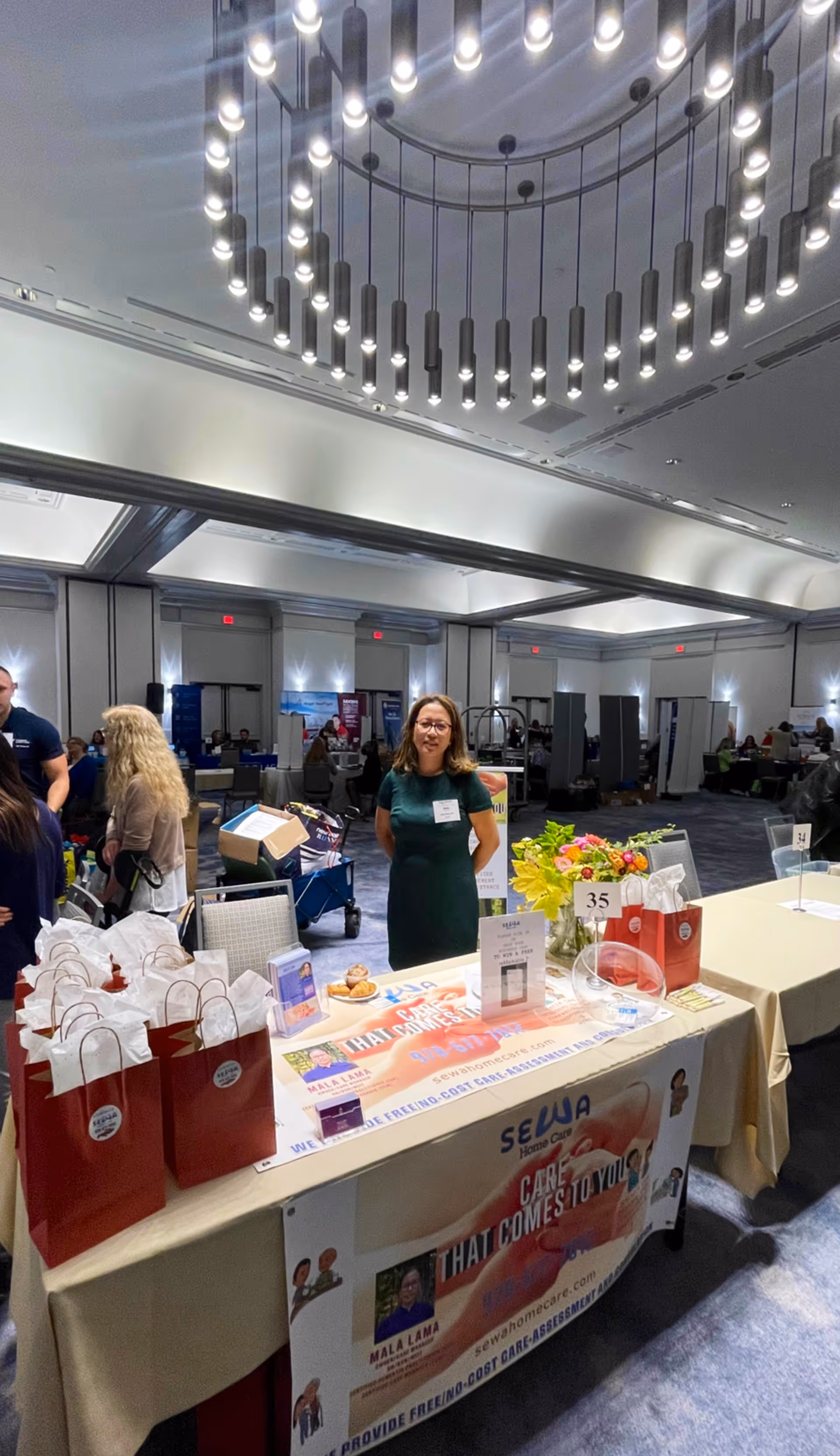 A woman stands behind a SEWA Home Care table at a bustling indoor expo booth.