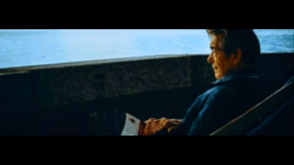 Elderly man sits in a chair on an outdoor deck by water.