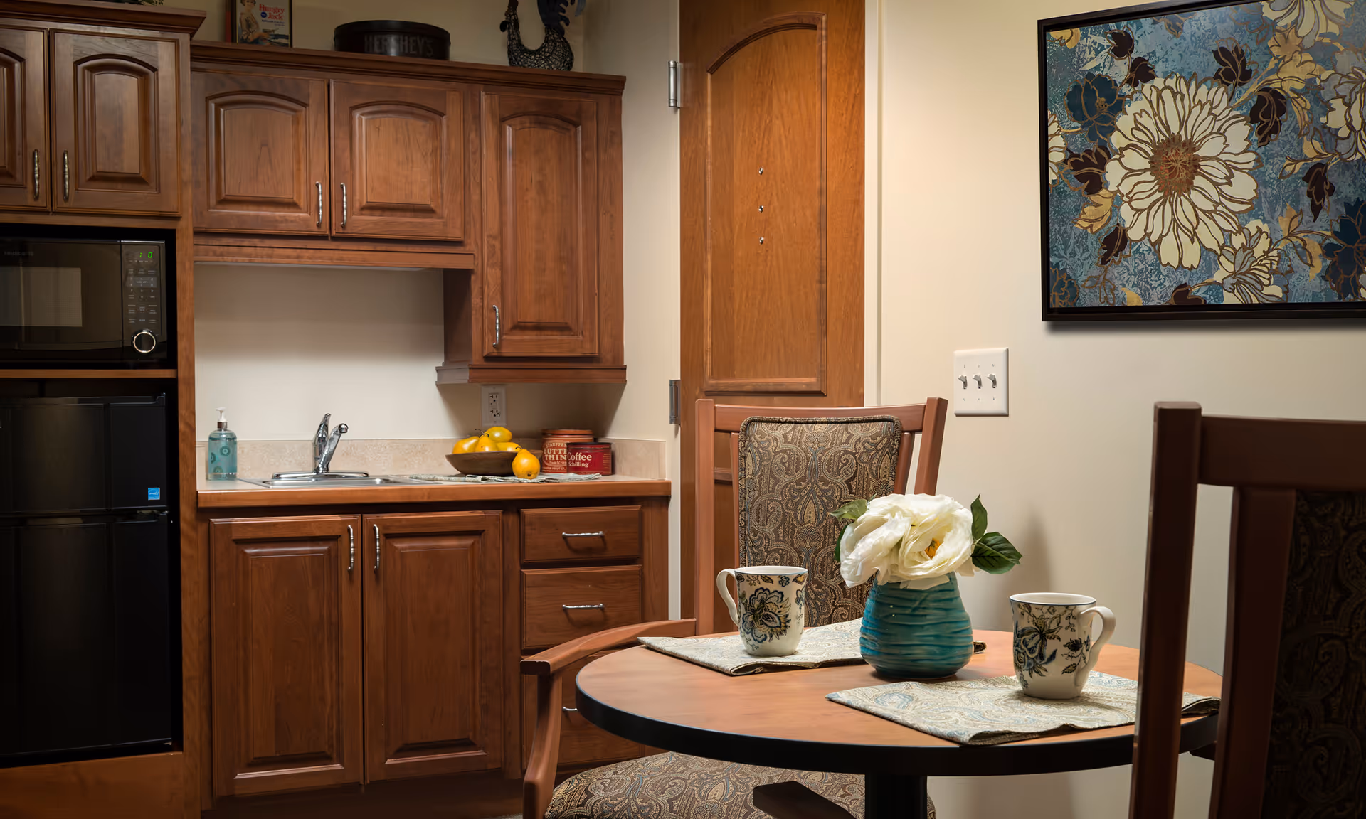 A cozy kitchen area with wooden cabinets, a small sink, a microwave, and a mini refrigerator. On the countertop, there is a bowl of lemons, a container of coffee, and a bottle of hand soap. In the foreground, there is a round wooden table with two patterned chairs, two floral mugs, and a blue vase with white flowers. A floral painting hangs on the wall.