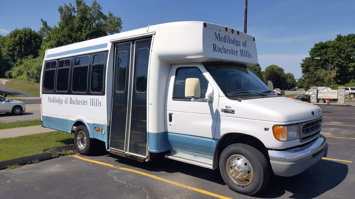 A white and blue shuttle bus parked in a parking lot with the text 'Medilodge of Rochester Hills' on the side and front. There are trees and a sign for Medilodge in the background.