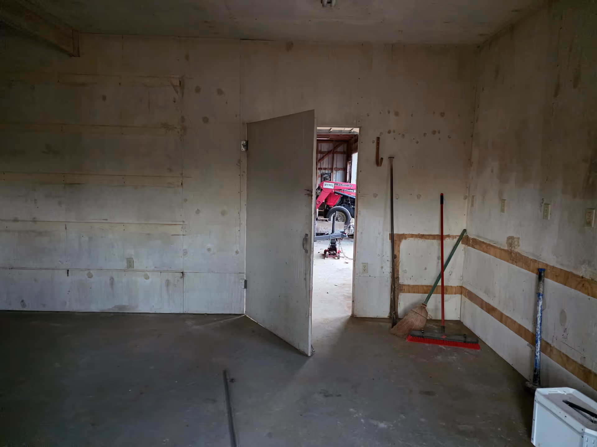 Unfinished interior room with concrete walls, doorway to outside; cleaning tools lean nearby.