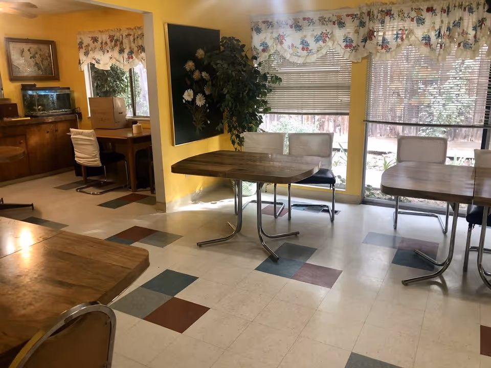 A bright dining area with yellow walls and large windows covered with floral valances. There are multiple wooden tables with metal legs and chairs with white backs and black seats. A large painting of white flowers hangs on the wall next to a tall green plant. In the background, there is a small desk with a chair, a fish tank on a wooden cabinet, and a window showing greenery outside.