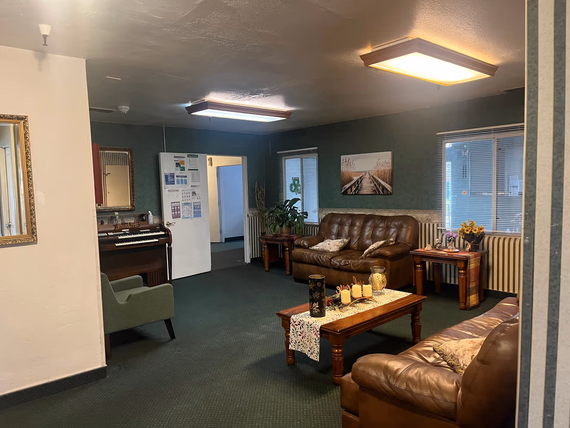 A cozy senior living common area with two brown leather sofas, a wooden coffee table decorated with candles and a vase, a green armchair, and a piano against the wall. The room has green carpet, green walls, two windows with blinds, framed artwork, and ceiling lights.