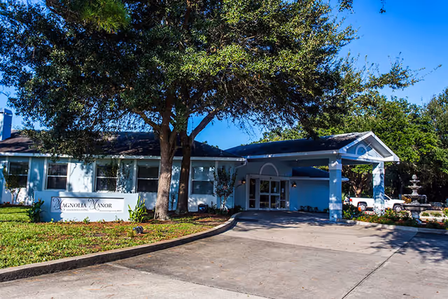 A single-story assisted living facility with a covered entrance, sign, and surrounding trees.
