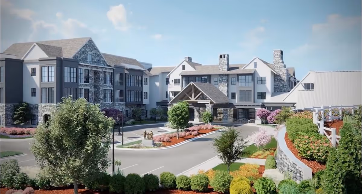 Exterior view of a senior living facility named Village Park Milton, showing a large multi-story building with stone and white siding, surrounded by landscaped gardens, trees, and a driveway with parking spaces.