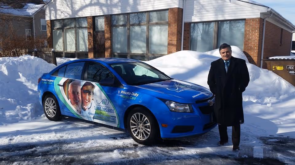 A man in a suit stands beside a blue A&D Home Health Solutions car in snow.