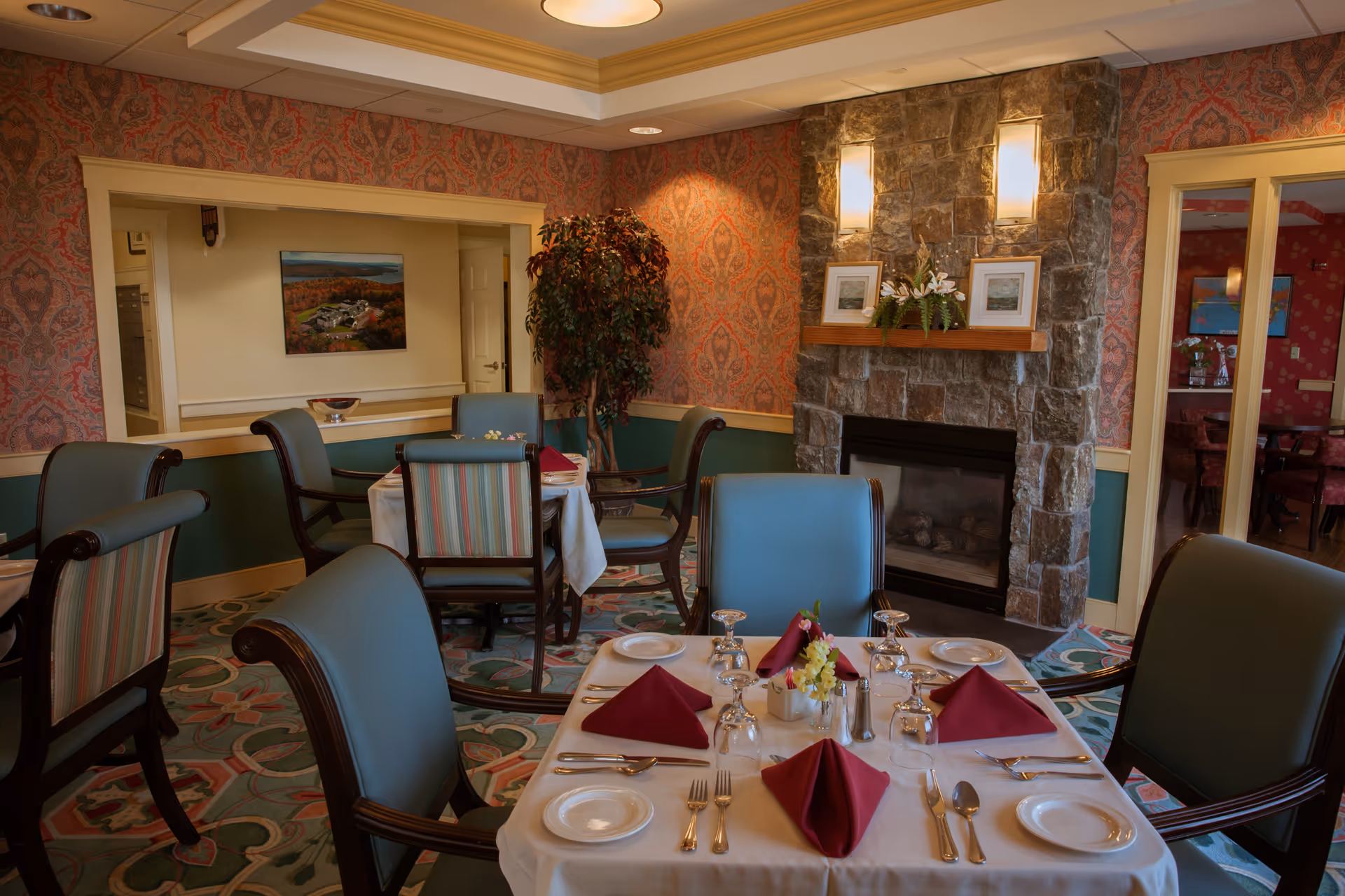 A formal dining room with a set table with burgundy napkins, upholstered chairs, patterned wallpaper, and a stone fireplace.