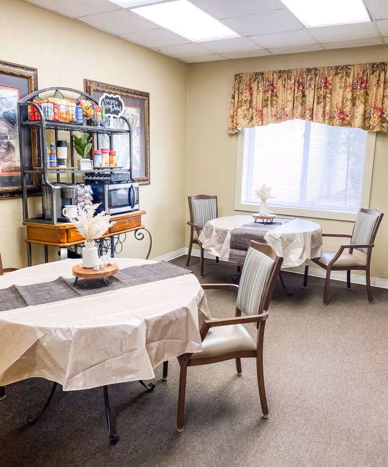 A small dining area with two round tables covered with beige tablecloths and dark table runners. Each table has a decorative centerpiece with dried flowers. There are wooden chairs with cushioned seats and striped backs around the tables. Against the wall, there is a black metal and wood shelving unit holding a microwave, coffee supplies, and snacks. The walls are light beige, and there is a window with floral valance curtains letting in natural light.