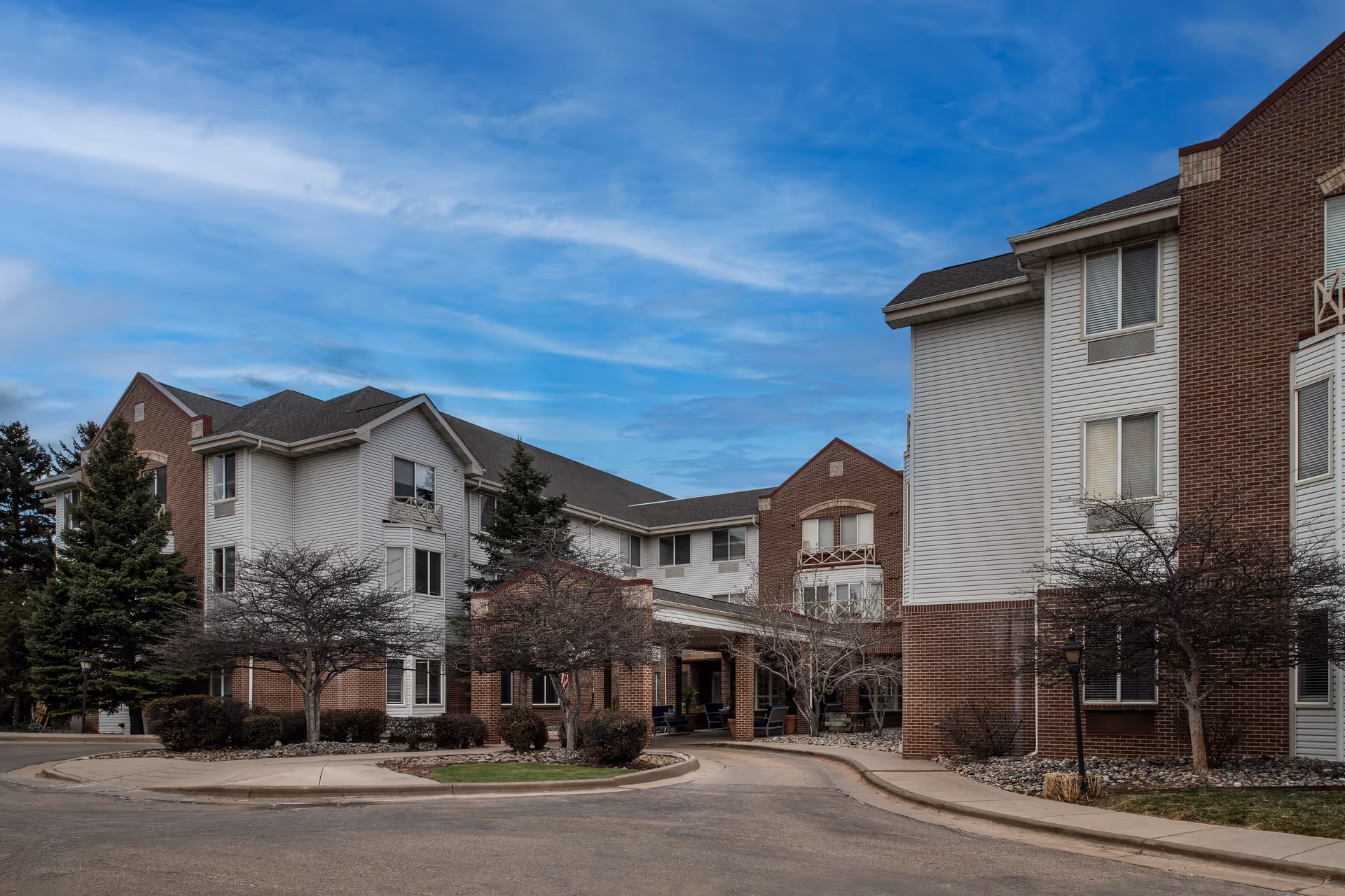 Exterior view of a multi-story assisted living facility building with brick and white siding, surrounded by trees and landscaping under a partly cloudy blue sky.