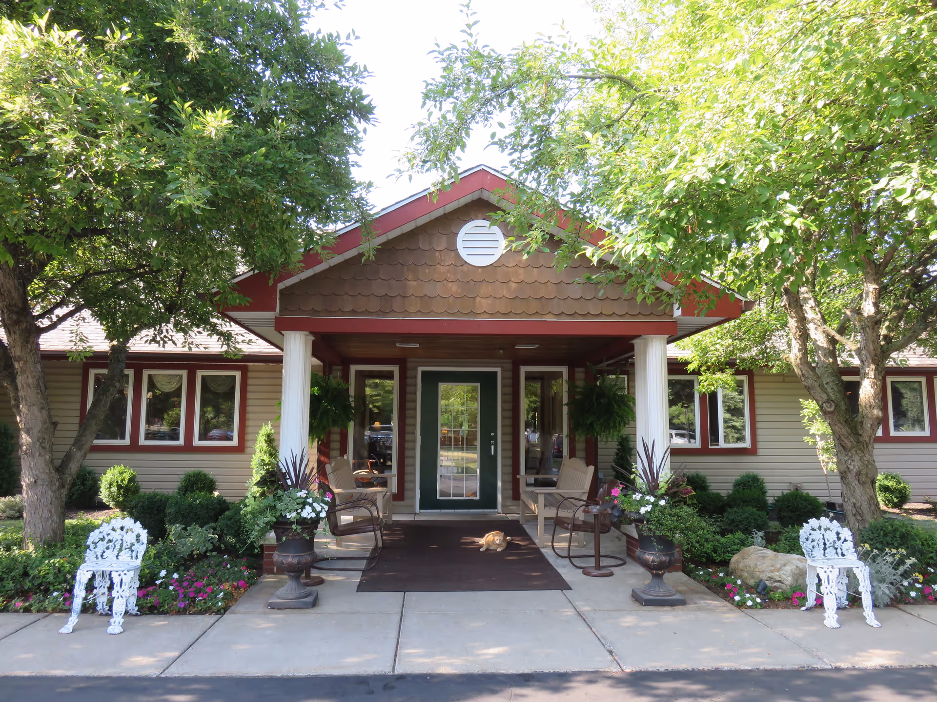 Front entrance of a single-story senior living facility with a covered porch supported by white columns. There are two white decorative chairs on either side of the entrance, surrounded by well-maintained flower beds and greenery. Trees provide shade over the pathway leading to the green door with glass panels. A small brown cat is sitting on the mat in front of the door.