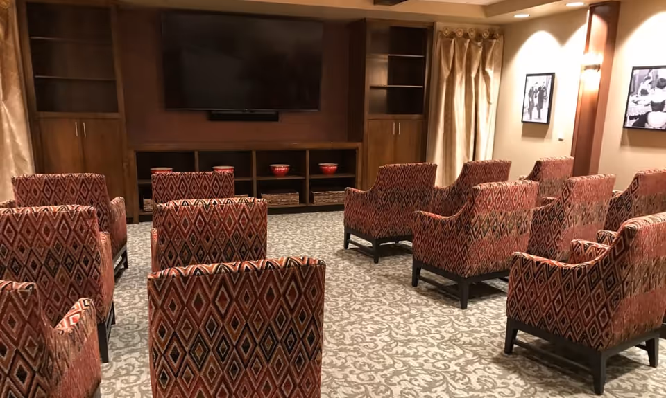 Small common room with patterned armchairs arranged in rows facing a wall-mounted TV and built-in wooden shelving.