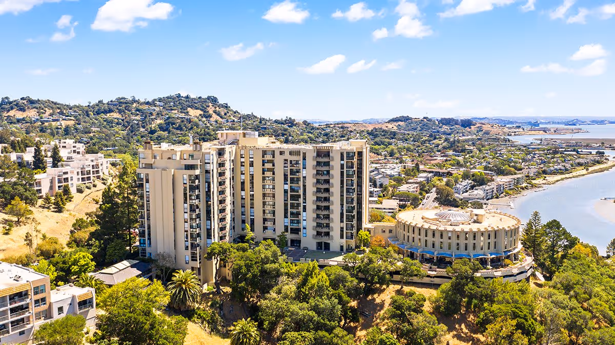 Aerial view of The Tamalpais Marin senior living facility showing a large multi-story building and a round building surrounded by trees, hills, and a body of water under a blue sky with scattered clouds.