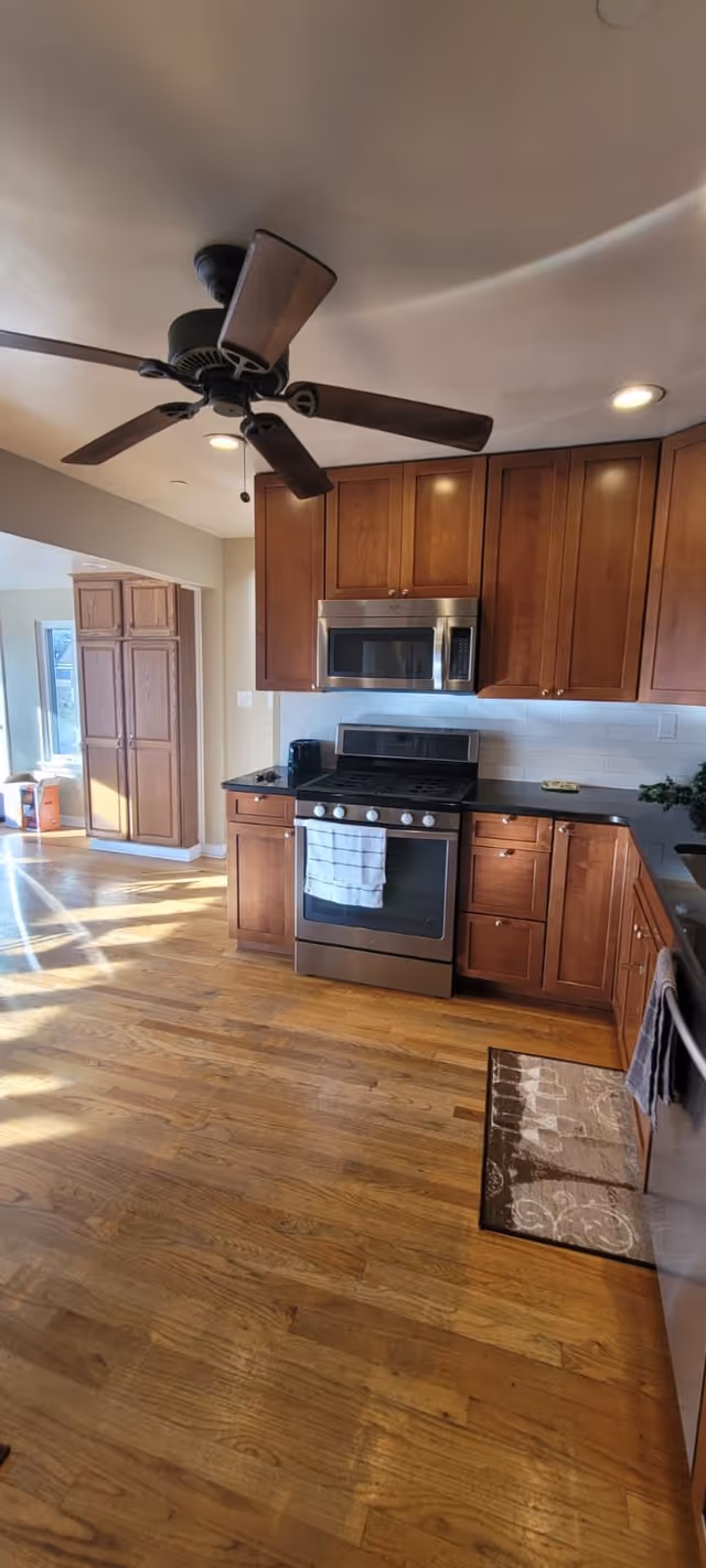 A kitchen with wooden cabinets, a stainless steel oven and microwave, a ceiling fan with wooden blades, hardwood floors, and a small rug in front of the sink area. Sunlight is streaming in from a window in the adjacent room.