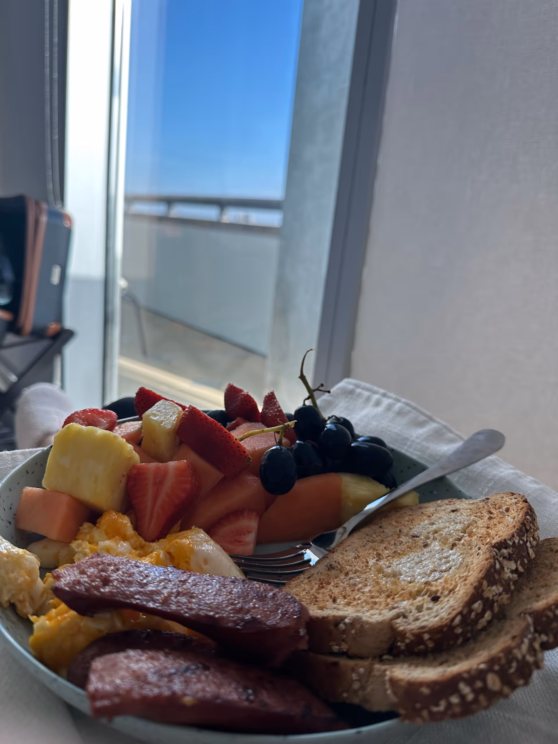 A plate of mixed fruit, scrambled eggs, sausage, and toast sits by a balcony window.