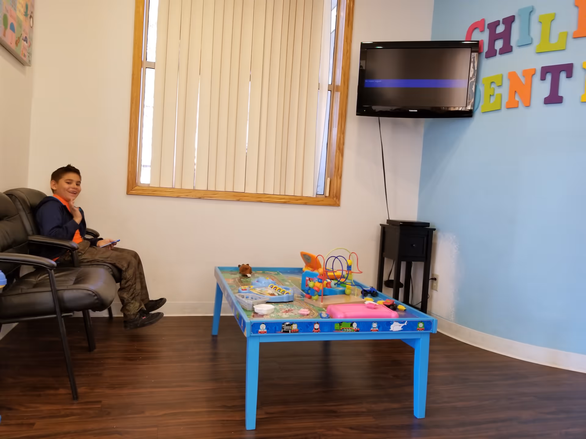 A child sits in a chair beside a blue toy table in a playroom.
