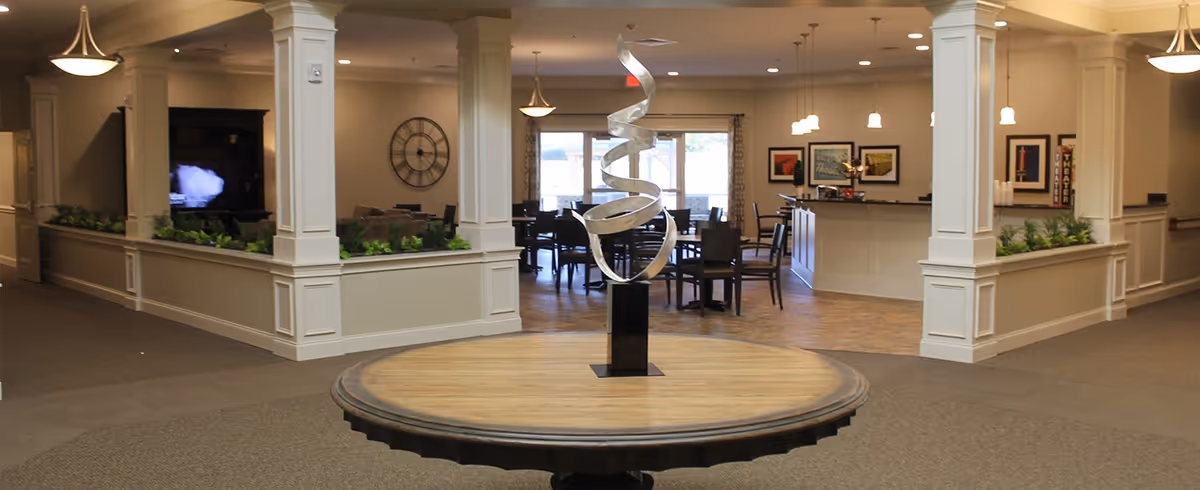 Interior view of a senior living facility showing a round wooden table with a modern spiral metal sculpture in the center. The background features a dining area with tables and chairs, decorative wall art, and pendant lighting. The space is open and well-lit with neutral tones and white columns.
