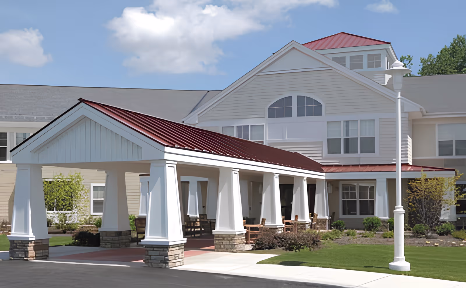 Front exterior of a senior living facility with a covered entrance supported by white columns, a red metal roof, and landscaped lawn.