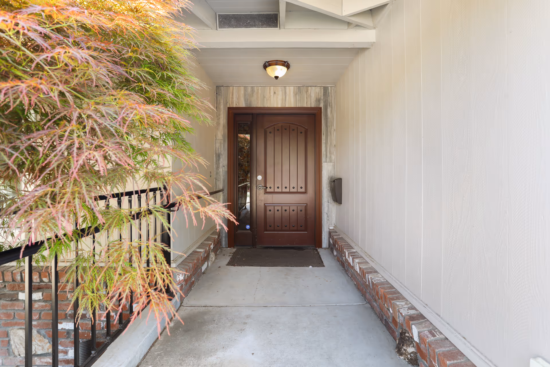 Covered front entry with a brown wooden door, brick low walls and foliage on the left.