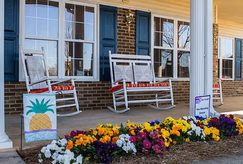 Front porch of a brick building with white columns, two white rocking chairs with red cushions, a colorful flower bed in front, and a small sign that reads 'The Manning House'.
