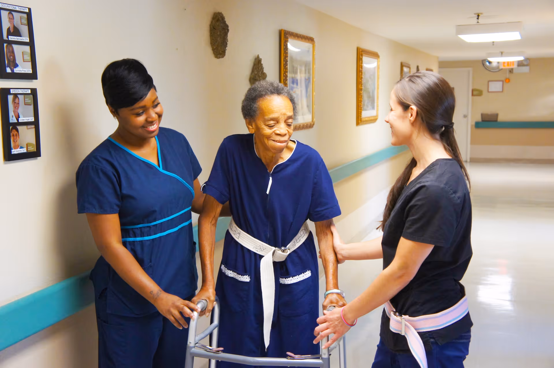 An elderly woman using a walker is being assisted by two female healthcare workers in a hallway of a senior living facility. The woman is smiling and wearing a navy blue outfit with a white belt, while the healthcare workers, dressed in scrubs, support her on either side. The hallway has framed pictures on the walls and a teal handrail.