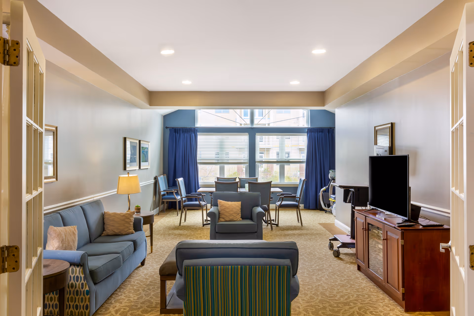 Cozy senior living lounge with blue seating, a TV, and windows with blue curtains.