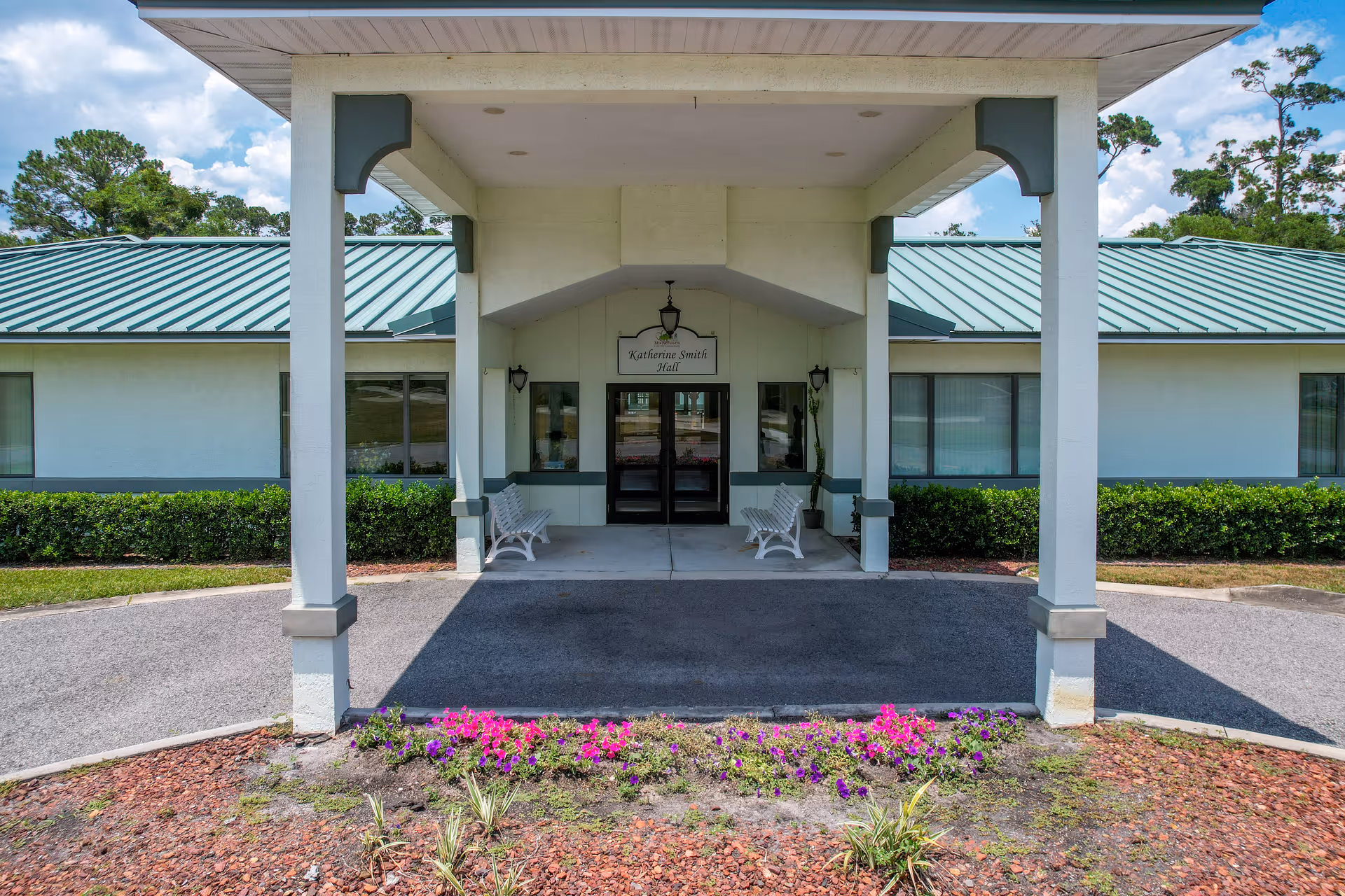Entrance of Katherine Smith Hall at Moosehaven, featuring a covered driveway with white pillars, two white benches on either side of the entrance, green bushes along the building, and a flower bed with pink and purple flowers in the foreground.