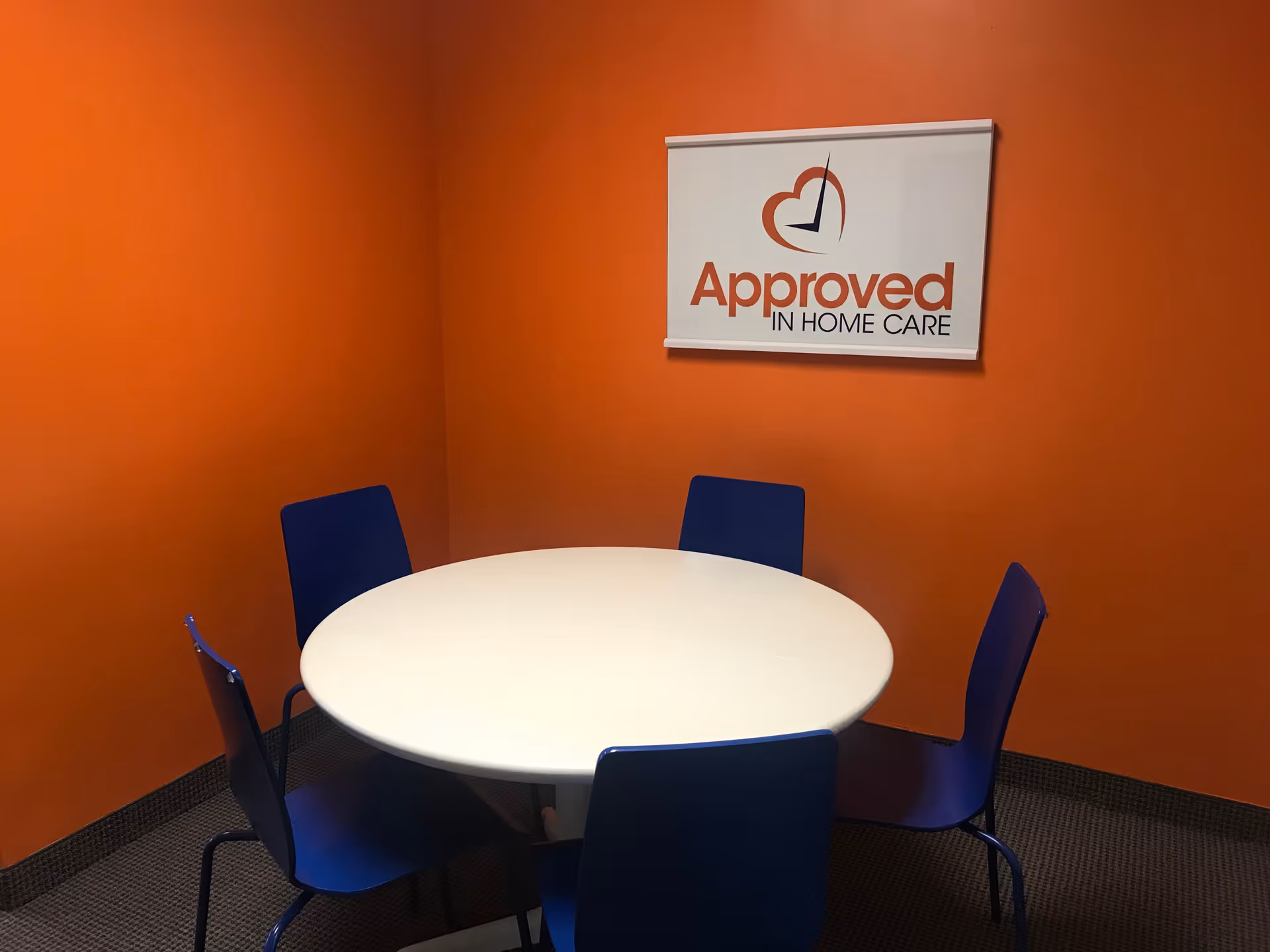 Orange-walled room with a round white table, blue chairs, and an Approved In Home Care sign.