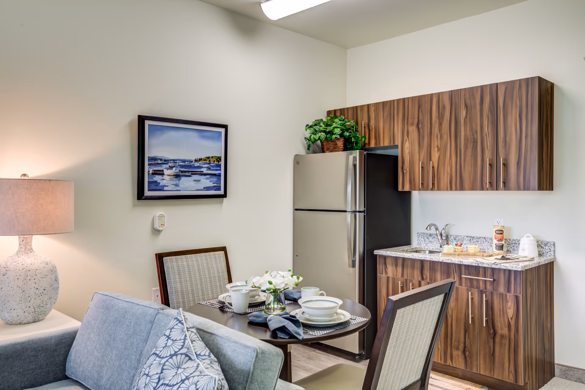 A small kitchen and dining area in a retirement living facility. The kitchen features wood-grain cabinets, a granite countertop with a sink, and a stainless steel refrigerator. A round dining table is set with white dishes, cups, and napkins, accompanied by two chairs. A gray couch with patterned pillows and a table lamp are partially visible. A framed painting of boats on water hangs on the wall above a thermostat. A potted plant sits on top of the refrigerator.