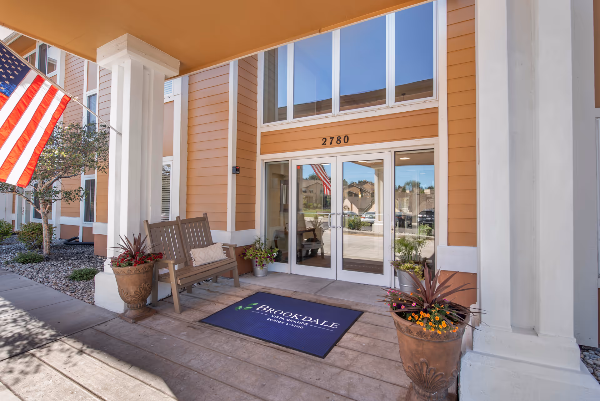 Exterior entrance of Brookdale Vista Grande with a bench, planters, and American flag.