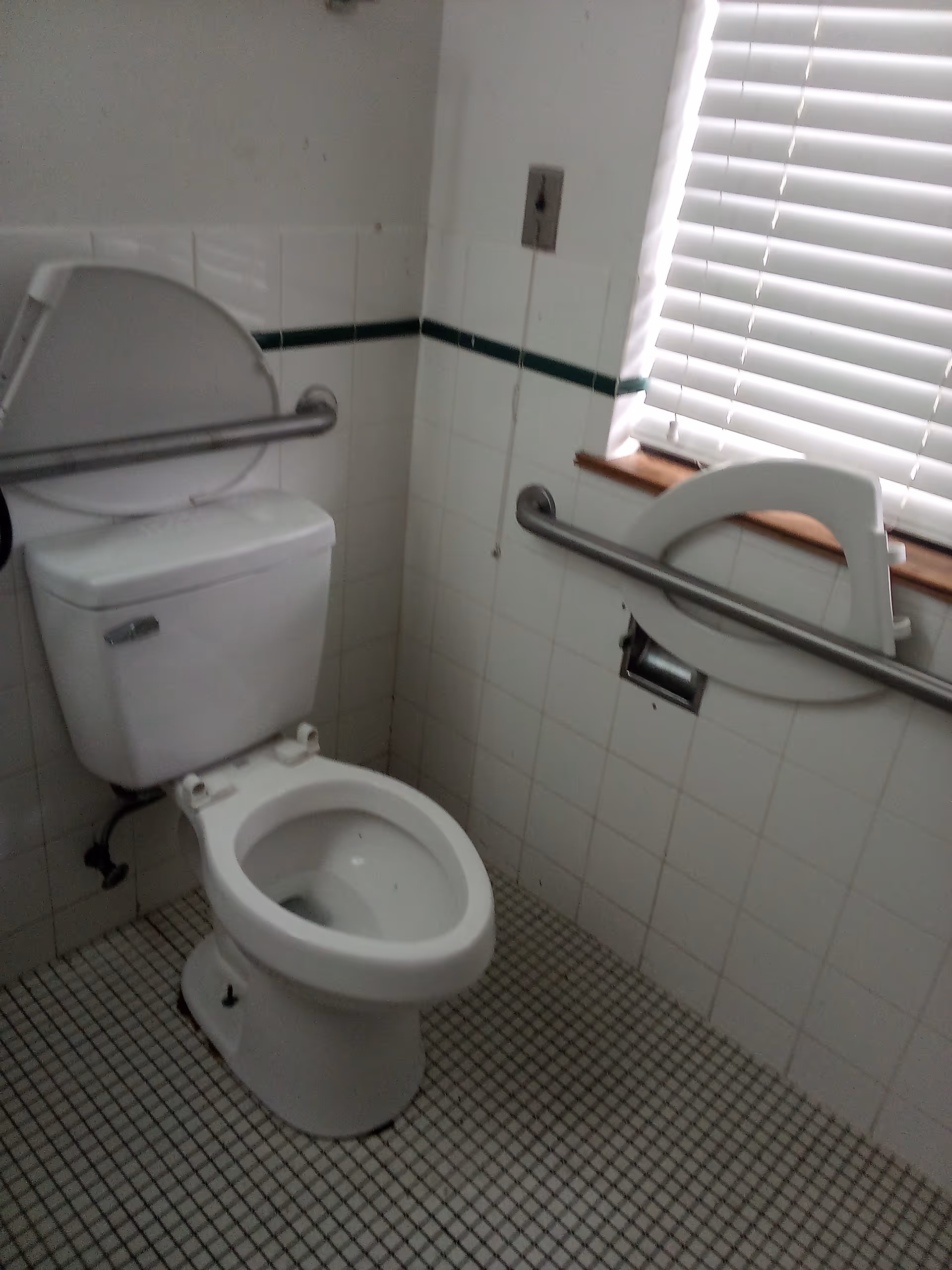 White bathroom with a toilet, wall grab bars, tiled walls, and a window with blinds.
