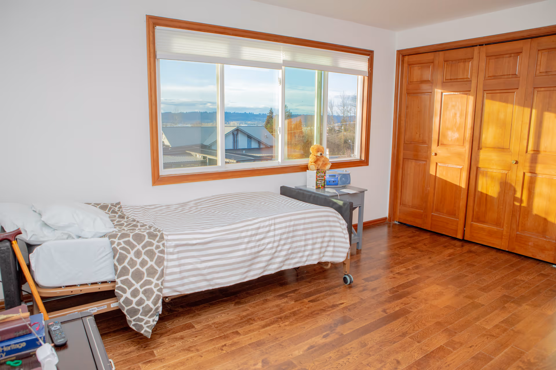 A cozy bedroom in a senior facility with a made bed, window, and wooden closet.
