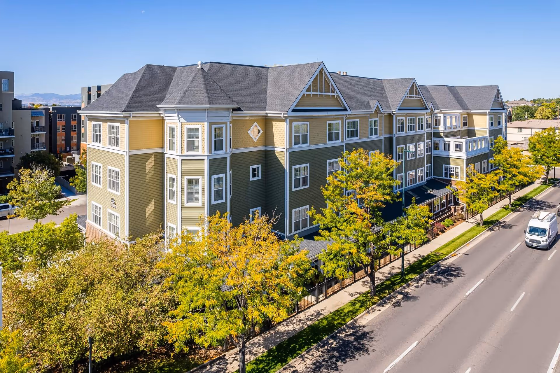 Exterior view of a multi-story senior living facility building with green and yellow siding, multiple windows, and a gray roof. The building is surrounded by trees with green and yellow leaves, and a road with a white van driving on it runs alongside the building.