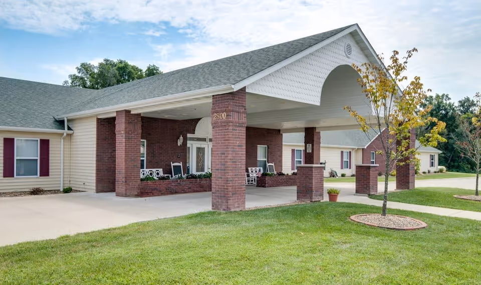 Exterior view of a single-story senior living facility with a covered entrance supported by brick columns. The building has beige siding, a gray shingled roof, and several windows with maroon shutters. There is a small tree and green lawn in the foreground under a partly cloudy sky.