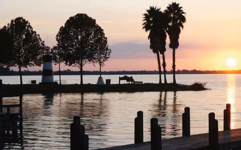 Sunset over a calm lake with palm trees, benches, and a lighthouse-like structure.